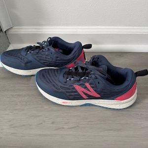 Könnte beinhalten: Paar marineblaue Sportschuhe mit roten Akzenten und weißen Sohlen. Die Schuhe haben ein strukturiertes Obermaterial, Schnürsenkel und ein rotes Markenlogo. Die Sohlen haben ein gesprenkeltes Design und einen blauen Akzent.