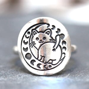May include: A silver ring with a cat sitting on a crescent moon with phases of the moon around it.