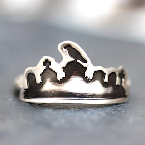 May include: A silver ring featuring a graveyard scene with a crow perched atop a tombstone. The ring has a dark, oxidized finish, and the design includes crosses and other details.