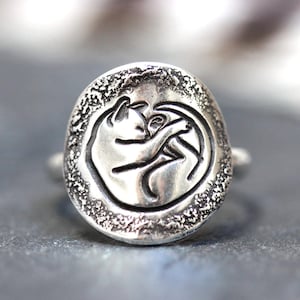 May include: A silver ring with a textured surface and a design of a curled up cat.