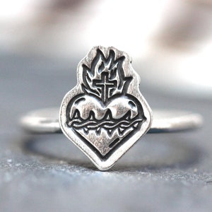 May include: A silver ring featuring a detailed design of a heart with a cross and flames. The heart is surrounded by thorns.