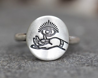May include: A silver ring with an oval-shaped design featuring a hand holding an eye with a sunburst radiating from it.