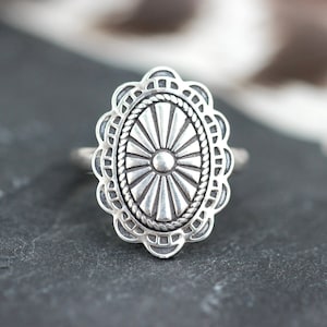 Puede incluir: Un anillo de plata con un diseño ovalado detallado que presenta un patrón de rayos de sol. El anillo tiene un borde texturizado y adornado.