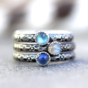 May include: Stack of three silver rings with intricate etched designs. Each ring features a small, round gemstone in shades of blue and white. The rings are stacked, showcasing the detailed craftsmanship.