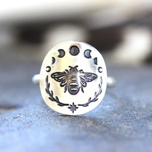 May include: A silver ring with a bee and moon phases design. The bee is in the center of the ring, surrounded by five moon phases.
