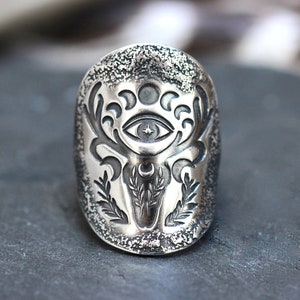 May include: A silver ring with a detailed design of a deer head with antlers, a crescent moon, and an eye with a star inside. The ring has a textured surface.