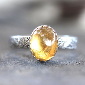 May include: A silver ring with a yellow oval gemstone set in a scalloped bezel. The band is decorated with a floral pattern.