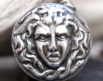May include: A silver pendant featuring a detailed relief of Medusa's face with snakes and wings. The pendant is round and has a loop at the top for attaching to a chain.