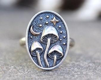May include: A silver ring featuring a mushroom design. The ring depicts three mushrooms with gold accents, a crescent moon, and a star. The ring is set against a dark blue background.
