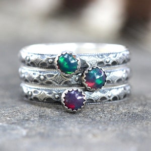May include: Three silver rings with opal gemstones. The rings are stacked on top of each other. The opals are green, red, and black.