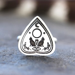 May include: A silver ring with a black and white ouija board design. The design features two black cats, a crescent moon, a circle, and an eye.