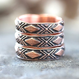 May include: Three stacked copper rings with a geometric pattern. The rings are a dark copper color with a slightly textured surface.