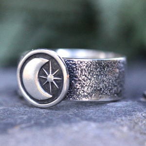 May include: A silver ring with a textured band and a circular design featuring a crescent moon and a star.