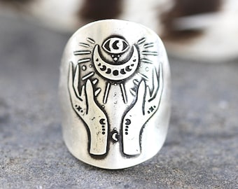May include: A silver ring with a design of two hands holding a crescent moon with an eye in the center. The moon is surrounded by a sunburst design.