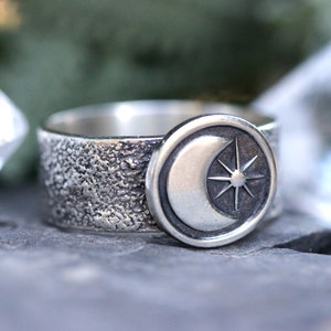 May include: A silver ring with a textured band and a circular design featuring a crescent moon and a star.