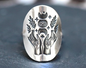 May include: A silver ring with an oval-shaped design featuring two hands holding a crescent moon, a circle with an eye, and a star. The design is surrounded by wheat stalks.