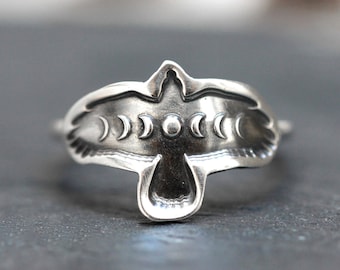 Puede incluir: Anillo plateado con un diseño estilizado de pájaro y fases lunares. El anillo presenta una silueta de pájaro con fases lunares en el centro. El anillo está hecho de un material metálico brillante.