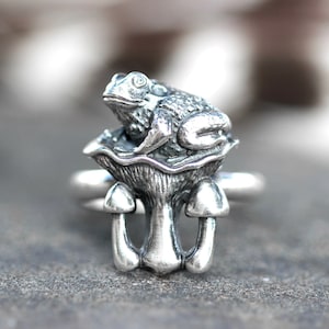 Puede incluir: Anillo plateado con un diseño detallado de una rana posada sobre una seta. El anillo tiene un diseño caprichoso, con la rana y la seta representadas en un estilo texturizado y tridimensional. La banda del anillo es simple y lisa.