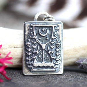 May include: A silver pendant with a detailed design of a crescent moon, stars, and a stylized tree with leaves. The pendant is rectangular and has a textured surface.