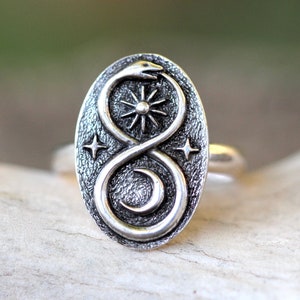 May include: A silver ring with an oval-shaped design featuring a snake curled into an infinity symbol. The snake is surrounded by a sun, moon, and stars.