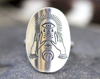 May include: Silver ring with an engraved design of two hands holding a crescent moon and an eye with a sunburst above it.