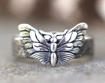 Könnte beinhalten: Ein silberner Ring mit einem detaillierten Schmetterlingsdesign. Die Flügel des Schmetterlings sind ausgebreitet und der Körper ist sichtbar. Der Ring ist aus strukturiertem Metall.
