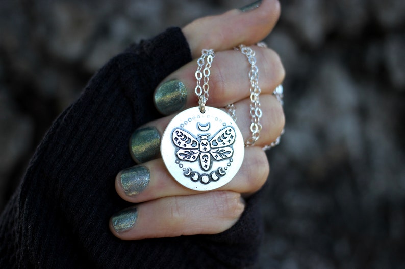 May include: A silver necklace with a round pendant featuring a detailed etched design of a moth with moon phases and stars.