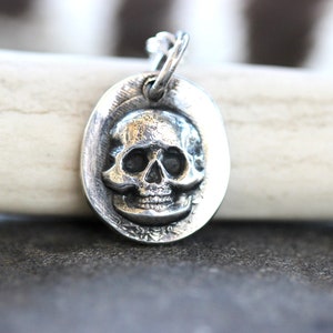 May include: A silver-colored pendant with a skull design. The skull is detailed with eye sockets, a nose cavity, and teeth. The pendant is oval-shaped and has a loop at the top for a chain. The background is a light-colored antler.