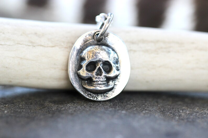 May include: A silver-colored pendant with a skull design. The skull is detailed with eye sockets, a nose cavity, and teeth. The pendant is oval-shaped and has a loop at the top for a chain. The background is a light-colored antler.