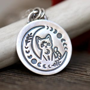 May include: A silver pendant necklace featuring a crescent moon with a cat sitting inside. The moon is surrounded by a circle of smaller moons and leaves.