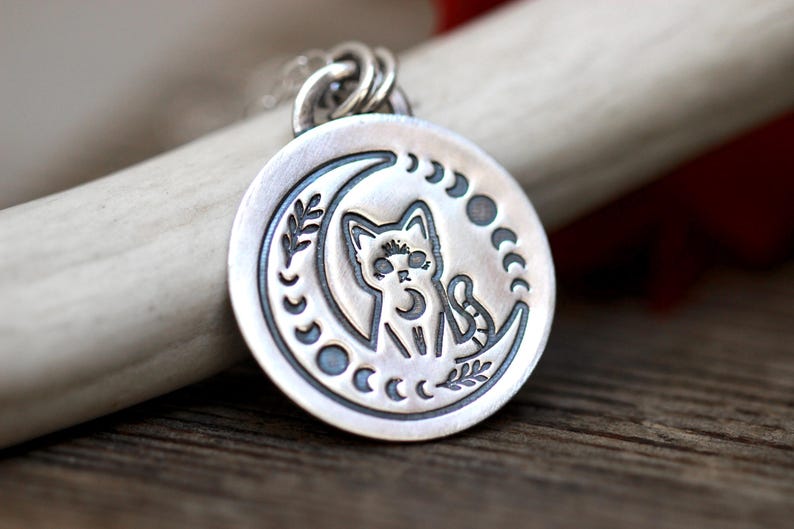 May include: A silver pendant necklace featuring a crescent moon with a cat sitting inside. The moon is surrounded by a circle of smaller moons and leaves.