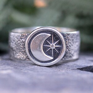 May include: A silver ring with a textured band and a circular design featuring a crescent moon and a star.