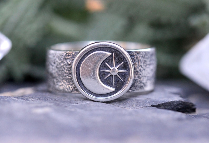 May include: A silver ring with a textured band and a circular design featuring a crescent moon and a star.