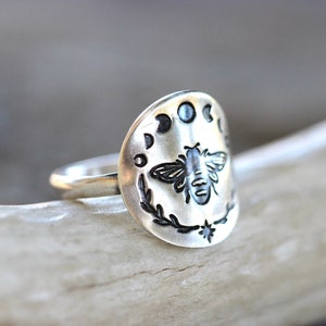 May include: A silver ring with a bee and moon phases design. The ring is engraved with a bee in the center, surrounded by a wreath and five moon phases.