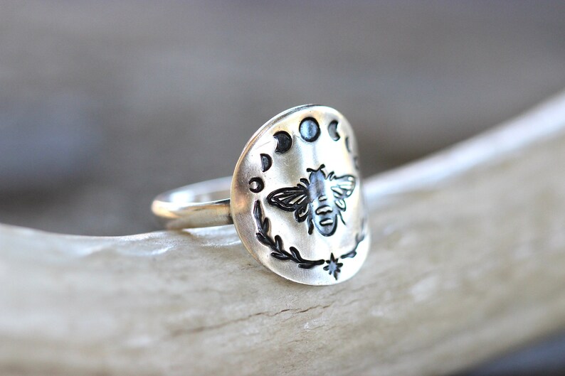 May include: A silver ring with a bee and moon phases design. The ring is engraved with a bee in the center, surrounded by a wreath and five moon phases.