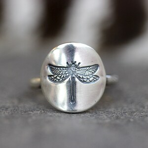 May include: A silver ring featuring a detailed dragonfly design. The dragonfly is centered on a smooth, oval-shaped face. The ring has a simple band and a slightly oxidized finish, giving it a vintage look.