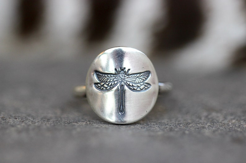 May include: A silver ring featuring a detailed dragonfly design. The dragonfly is centered on a smooth, oval-shaped face. The ring has a simple band and a slightly oxidized finish, giving it a vintage look.