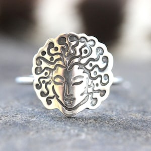 May include: A silver ring with a detailed face design and flowing hair. The face is set within a circular frame with a decorative edge. The ring is shown against a blurred background.
