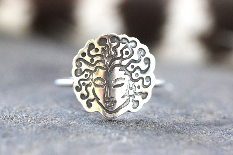 May include: A silver ring with a detailed face design and flowing hair. The face is set within a circular frame with a decorative edge. The ring is shown against a blurred background.