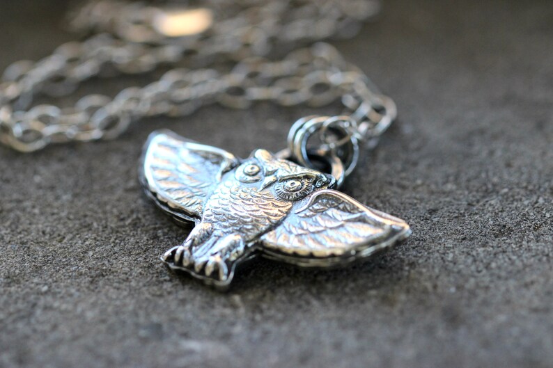 May include: Silver owl pendant necklace with a detailed design. The owl is depicted with its wings spread, and its eyes are large and round. The necklace is made of a silver chain.