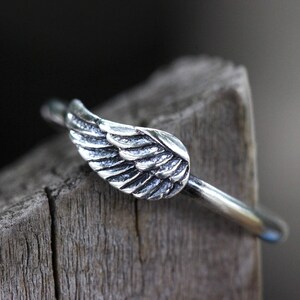 May include: A silver ring with a wing design. The ring is on a wooden surface.