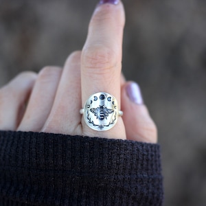 May include: A silver ring with a bee design surrounded by a circle of moons. The bee is black and the moons are silver with black centers.