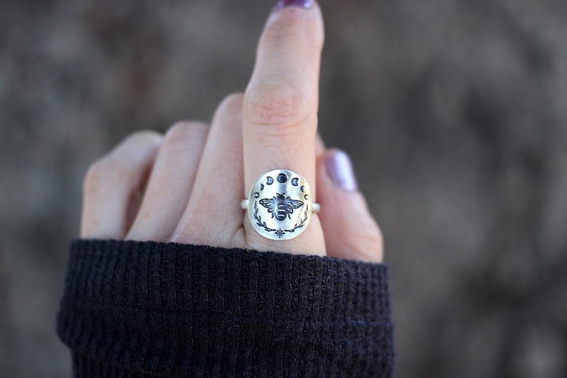 May include: A silver ring with a bee design surrounded by a circle of moons. The bee is black and the moons are silver with black centers.