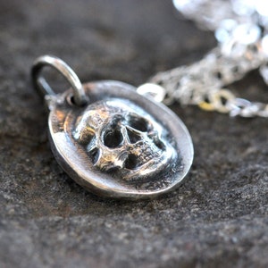 May include: A silver pendant necklace featuring a detailed skull design. The skull is set within a circular frame, attached to a silver chain. The pendant is made of a shiny, metallic material.