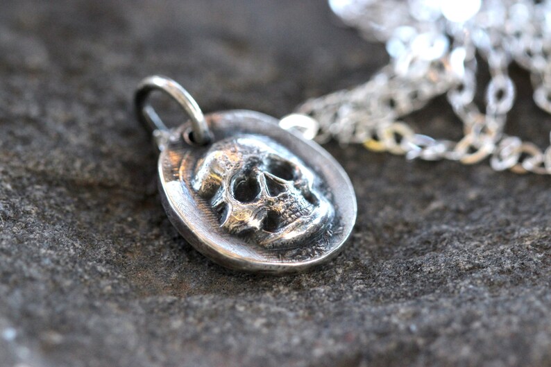 May include: A silver pendant necklace featuring a detailed skull design. The skull is set within a circular frame, attached to a silver chain. The pendant is made of a shiny, metallic material.