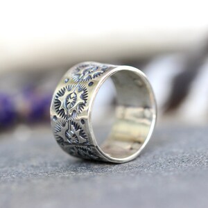 May include: A wide, silver ring with a detailed pattern of repeating starburst and gear-like designs. The ring is displayed on a gray surface, with a blurred background of purple flowers and white feathers.
