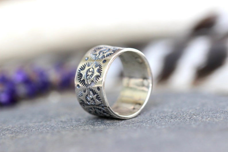 May include: A wide, silver ring with a detailed pattern of repeating starburst and gear-like designs. The ring is displayed on a gray surface, with a blurred background of purple flowers and white feathers.