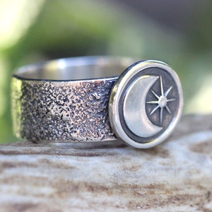 May include: A silver ring with a textured band and a circular design featuring a crescent moon and a star.