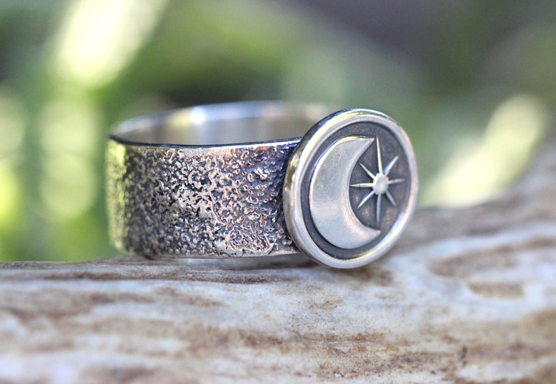 May include: A silver ring with a textured band and a circular design featuring a crescent moon and a star.
