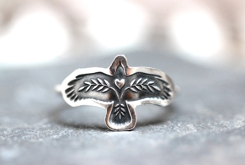 May include: A silver ring featuring a detailed bird design with outstretched wings. The bird has a heart and leaf details. The ring is set against a gray background, showcasing the intricate craftsmanship of the jewelry.